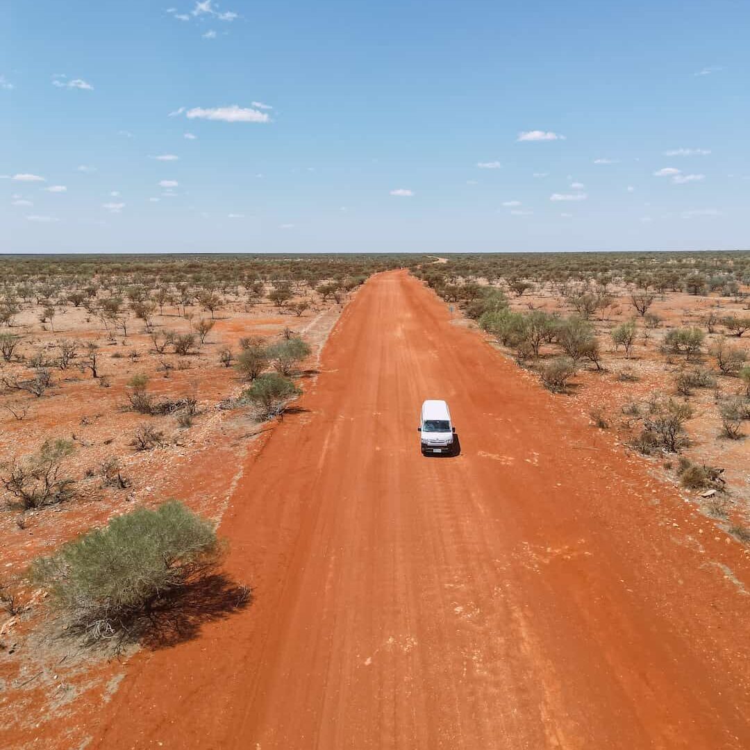 Drone shot from sky of vast outback WA road with red dirt, and a booked 2-3 seater high top campervan